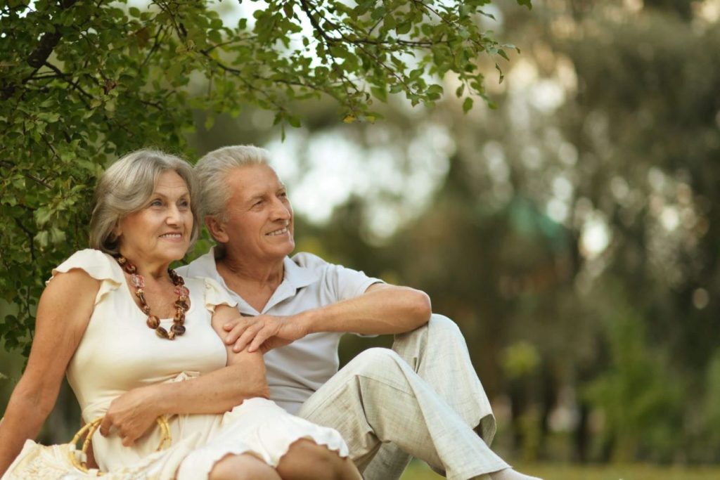 An elderly couple sits together outdoors, smiling and looking into the distance. Under a tree with lush green leaves, they savor a tranquil moment in the park. With gray hair and her in a beige dress, this scene reflects the serene lifestyle championed by Fortuna elder care homes.